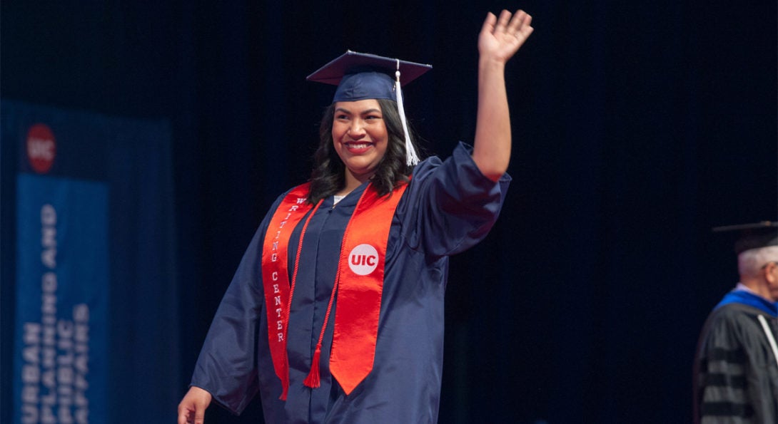 A woman wearing a cap and gown and red sash waves at graduation