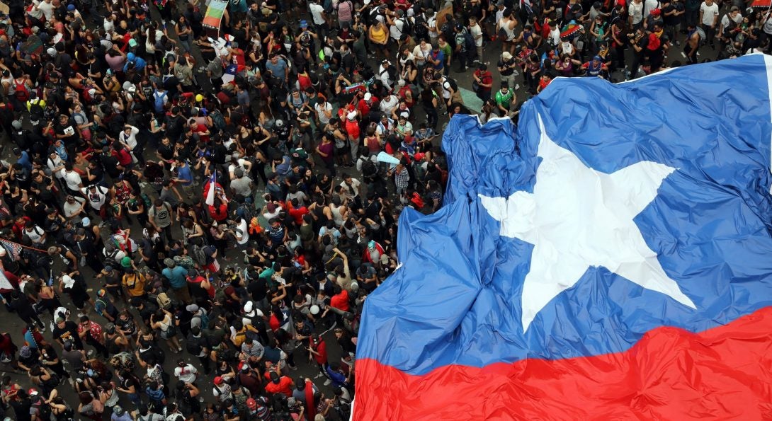 A crowd of protesters are seen from above, partially covered by an enormous Chilean flag