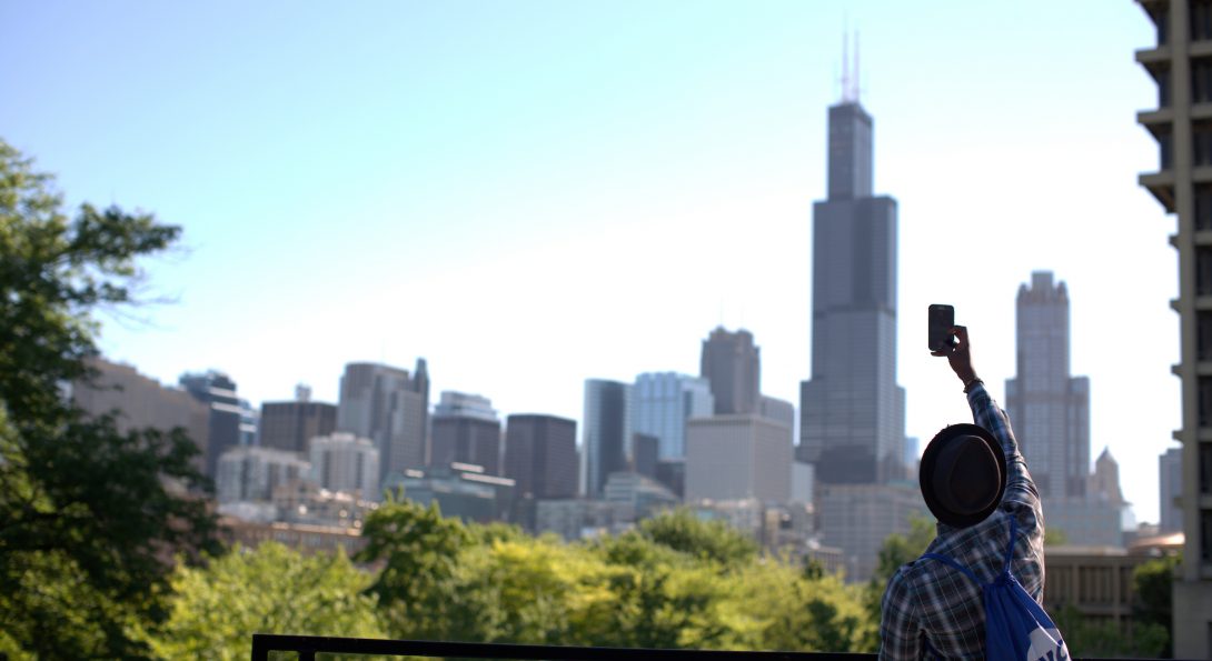 A young person in a hat takes a long distance photo of Willis Tower from UIC's campus.