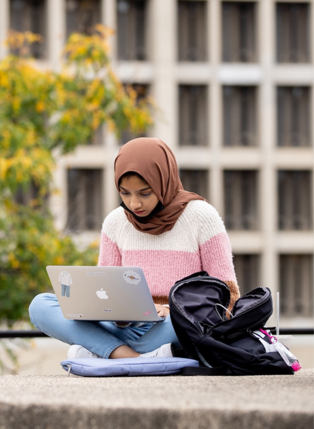 A woman in a hijab is sitting cross legged with her laptop on her lap and backpack by her side.