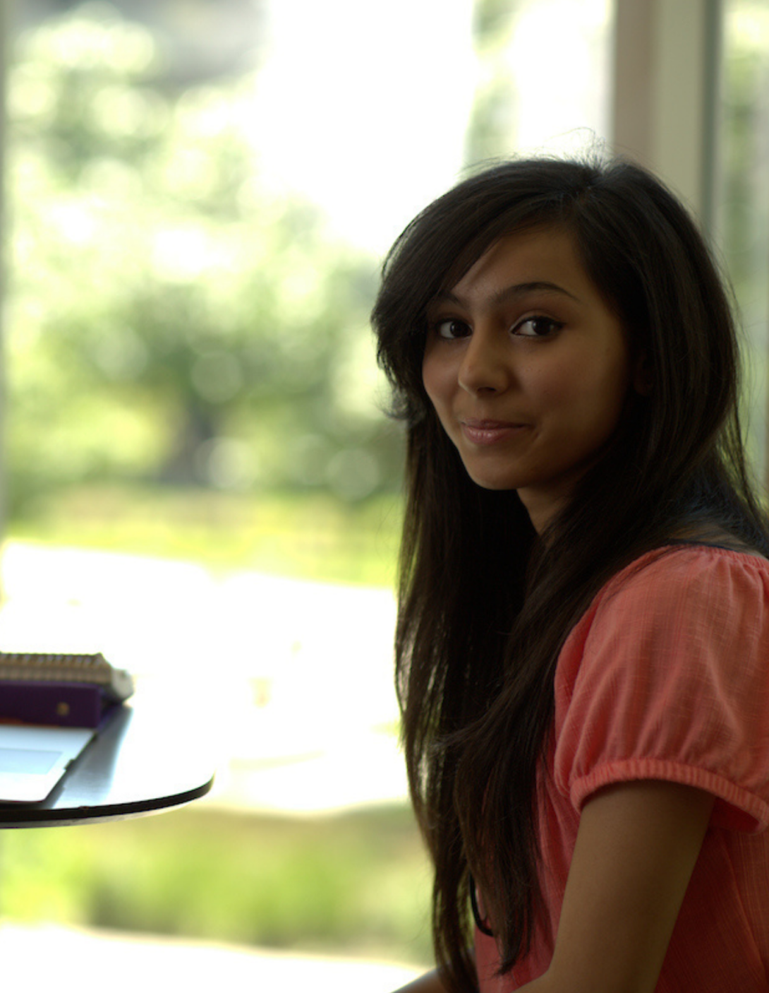 female student staring into camera over a blurred, sunny window backdrop. she has long black hair and a coral blouse, and is smirking.